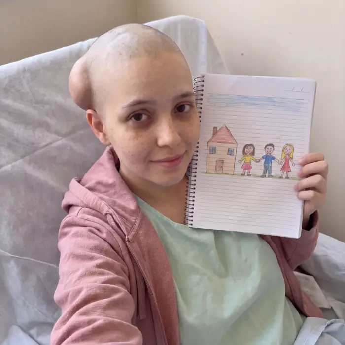 Ela na cama do hospital, de cabeça raspada, segurando um caderno com o desenho de uma casa e uma família